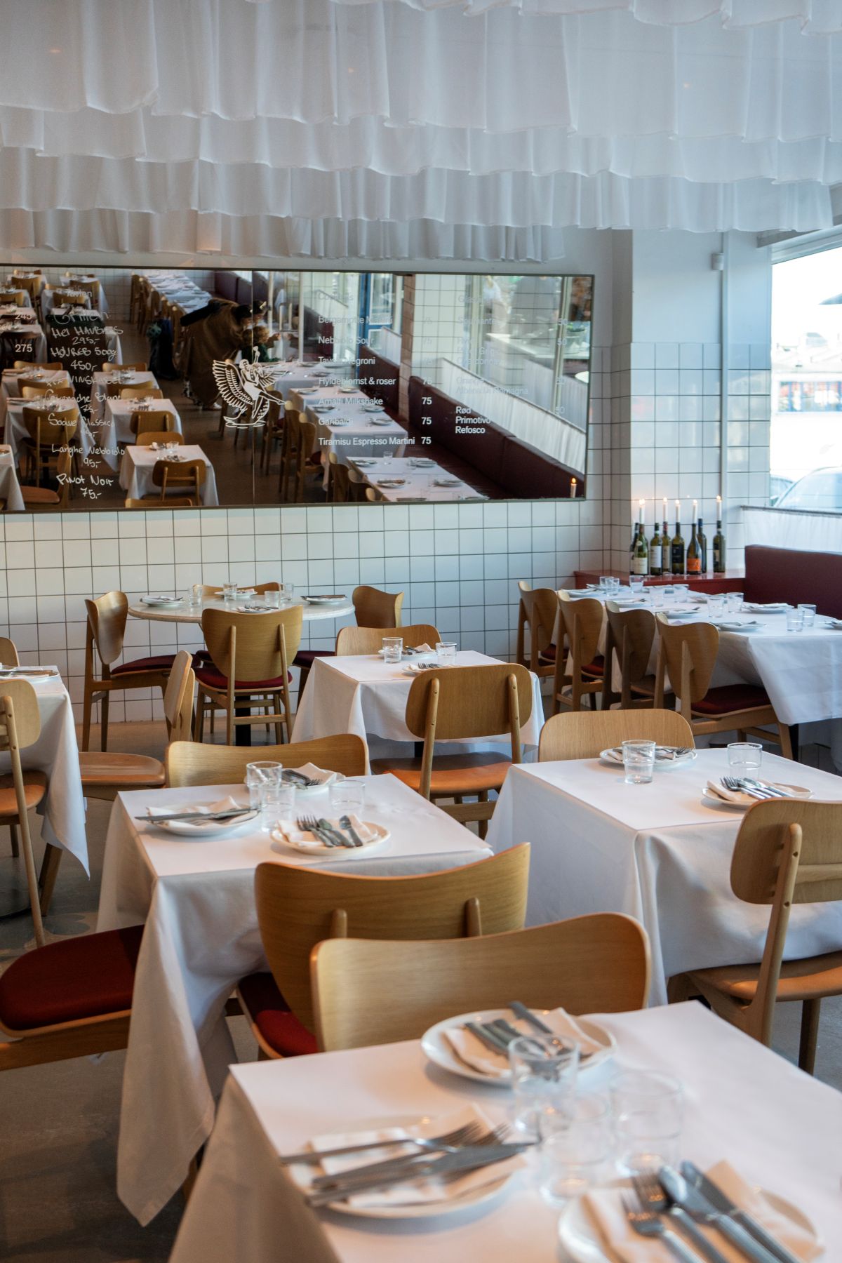 A bistro restaurant dining room with wooden chair, white tablecloths, silver cutlery, white plates, tiled walls, and white ceiling drapery.