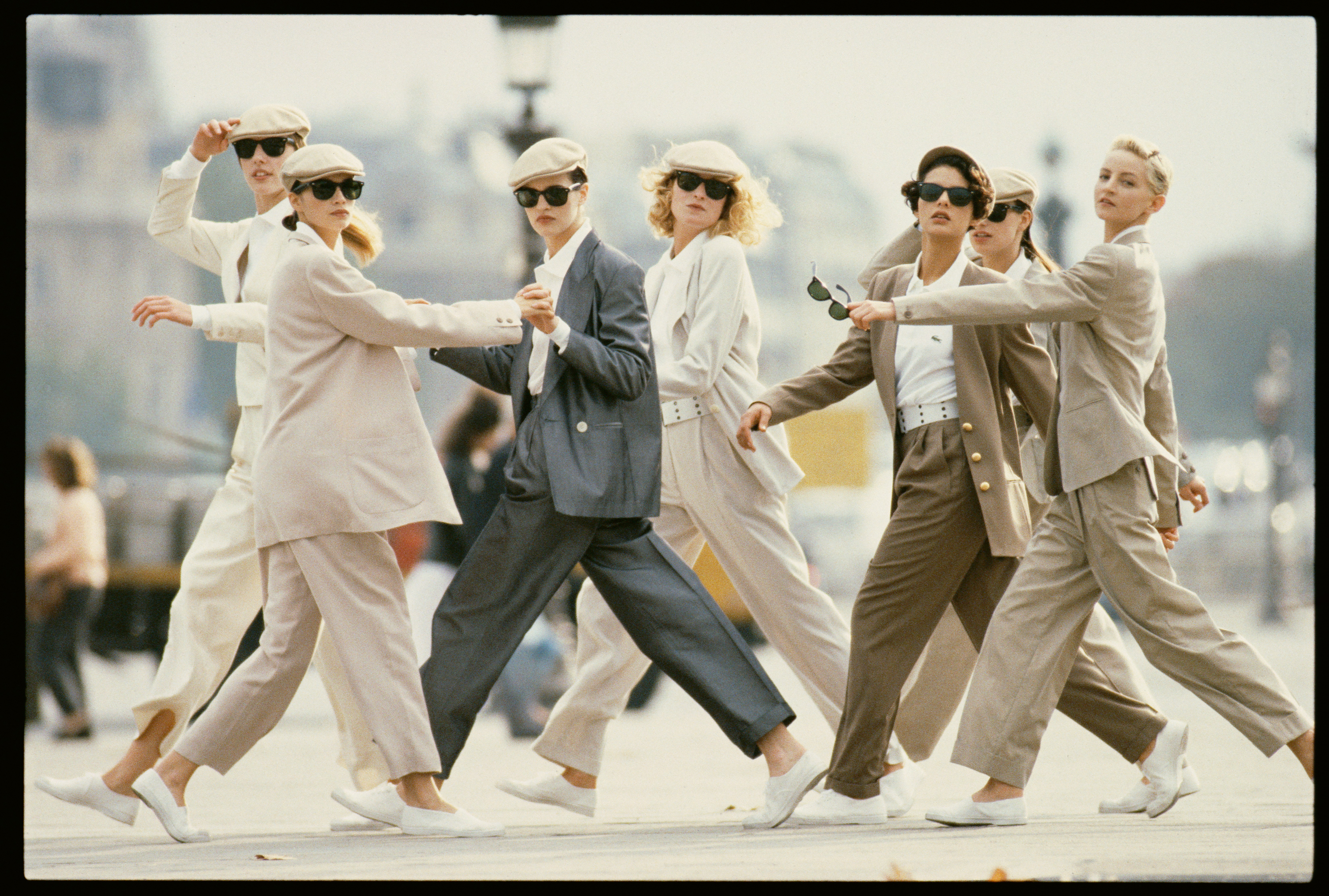 Seven models in beige and grey tailoring, sunglasses and flat caps stride together across a city square in a tight, dynamic pack.