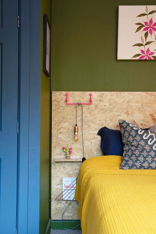A bedroom with olive green wall paint, OSB headboard, framed floral wall art, floating shelf and magazine rack fixed to wall