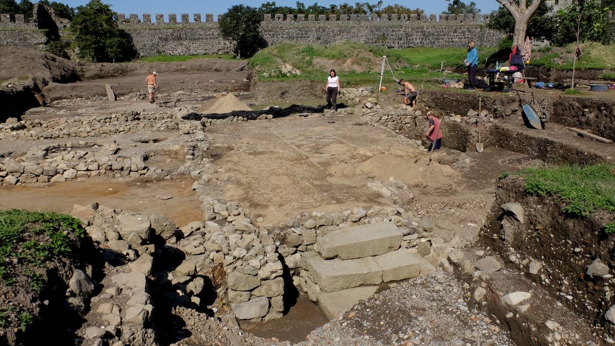 We see an archaeological site with different levels of dirt and stone. There are five people excavating at the site, which has a stone wall in the background.