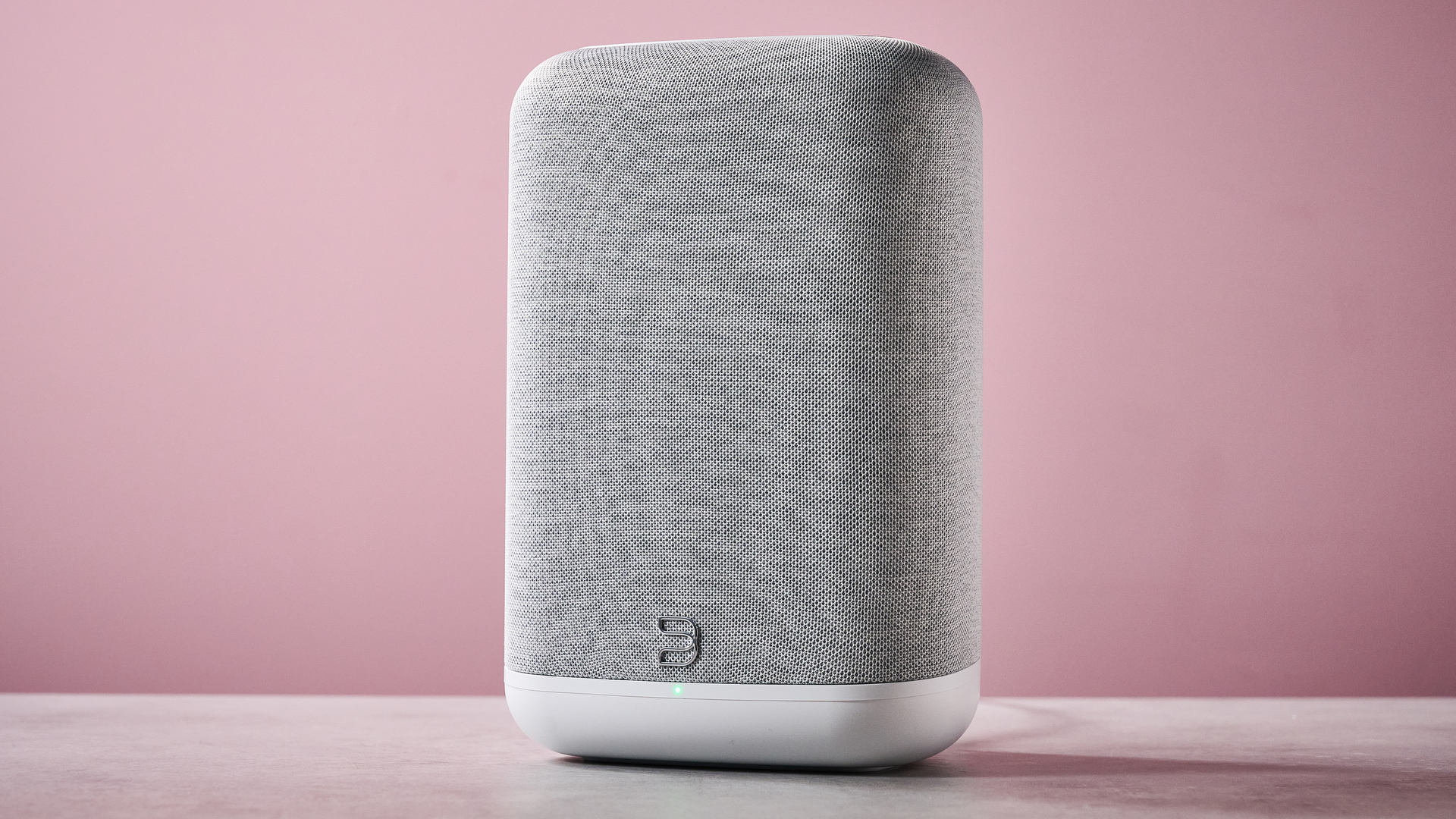 The Bluesound Pulse Flex at a 3/4 angle on a stone effect counter in front of a pink background.