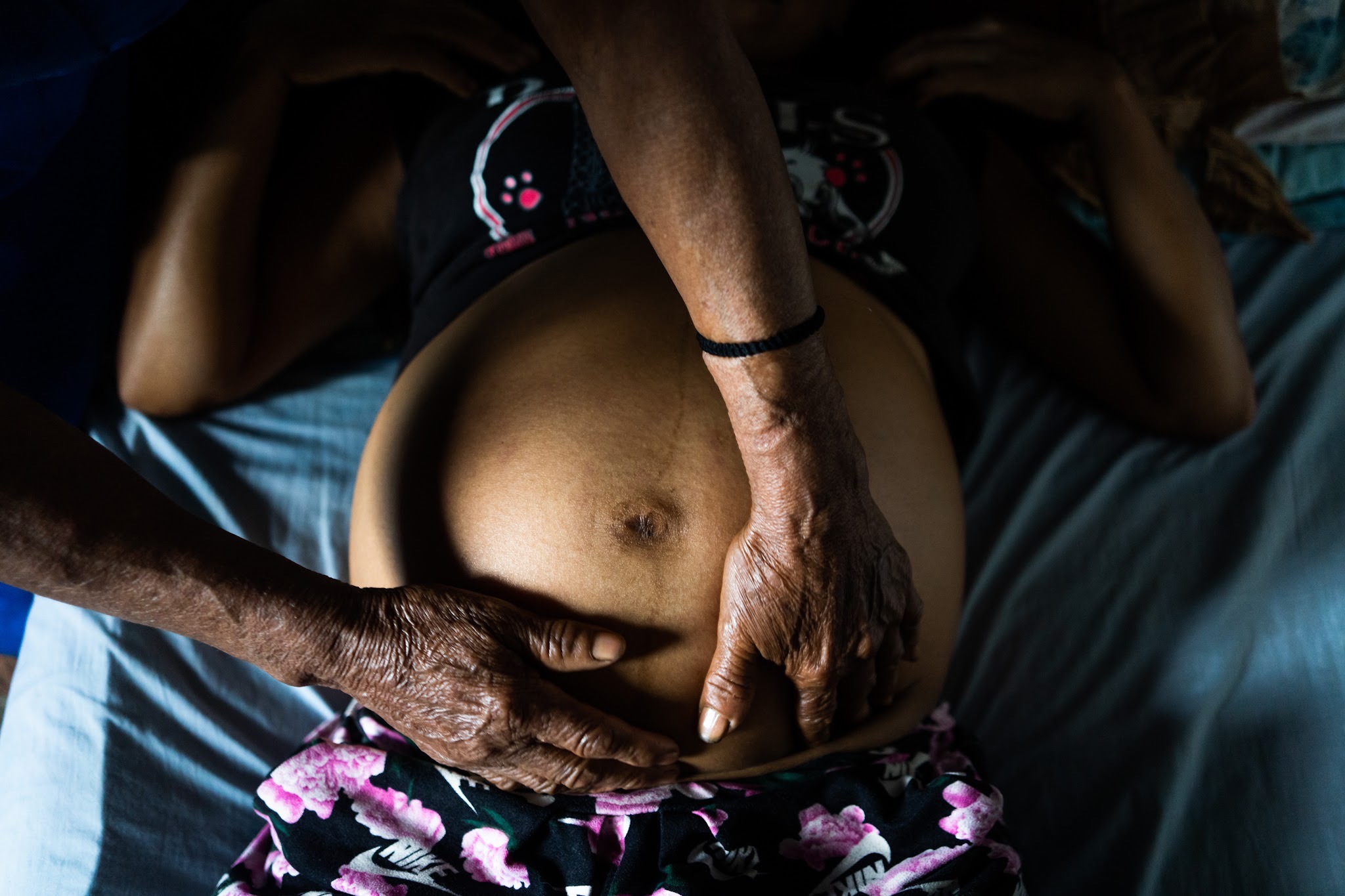An overhead shot of two pairs of weathered hands pressing gently on a pregnant woman's bare belly as she lies on a bed.