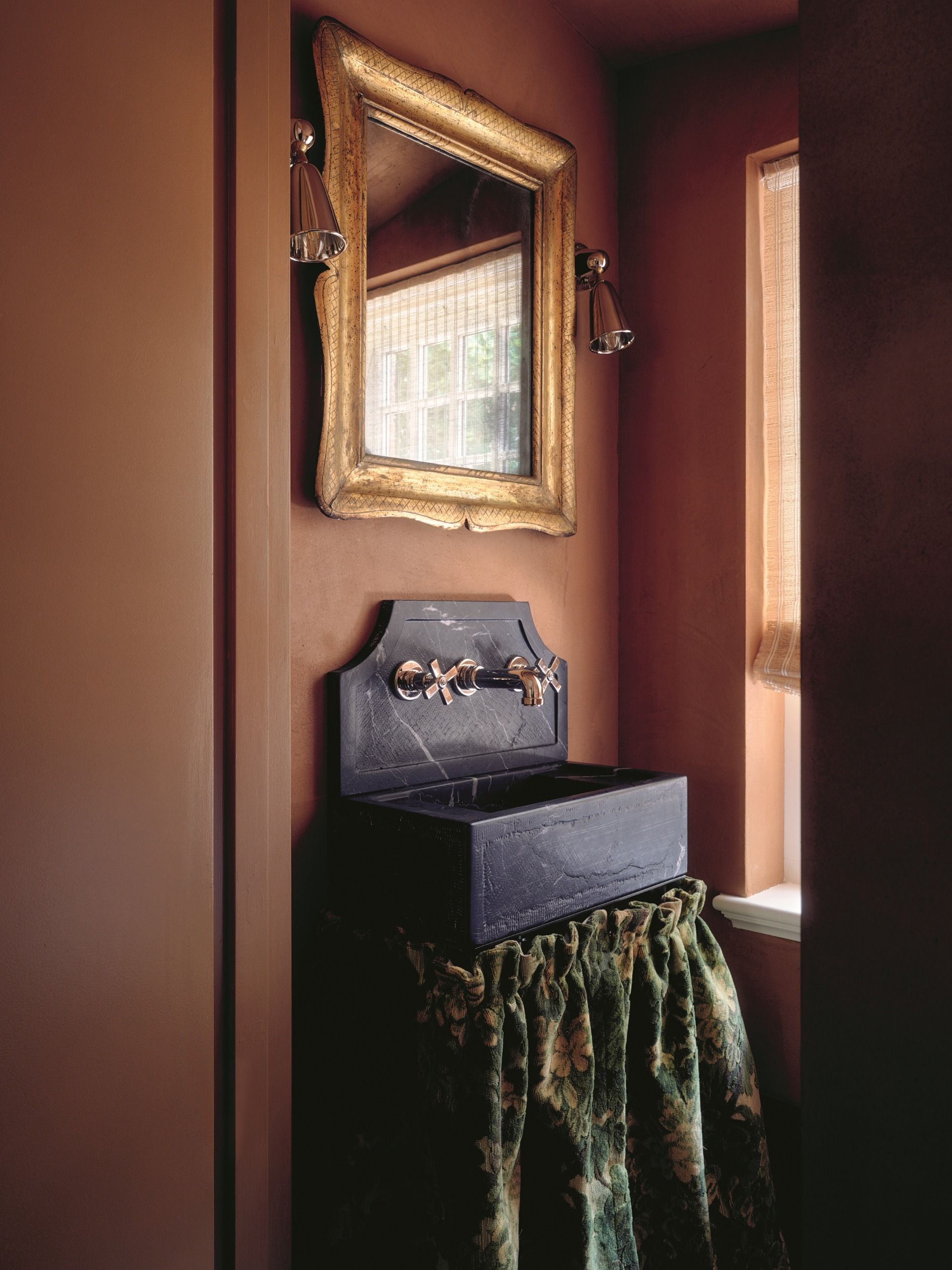 a terracotta bathroom with a black sink and sink skirt, with a gilded mirror 