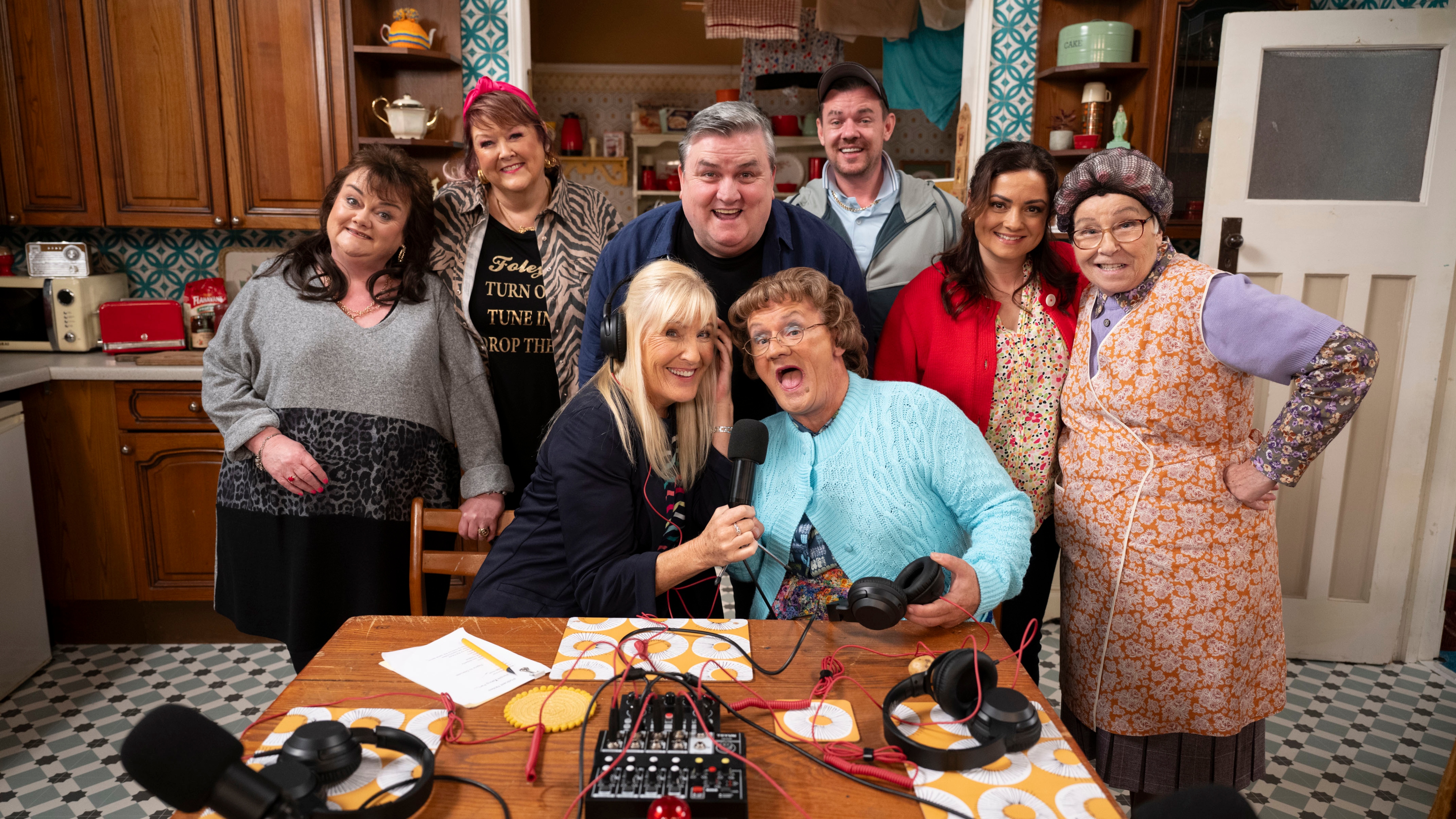 Mrs Brown and daughter Cathy sitting at the kitchen table at a pod cast recording set up with their friends and neighbours surrounding them.