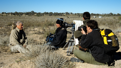 David Attenborough with a meerkat on his shoulder during filming