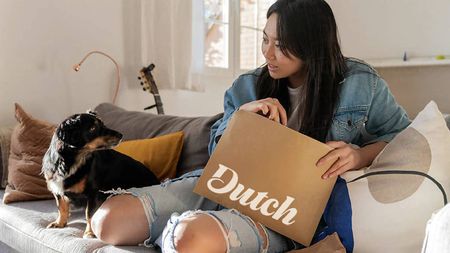 A woman holding a parcel from Dutch Pet Inc. with her dog, sat on the sofa.