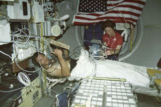 Astronauts Bonnie Dunbar and Larry DeLucas conduct a blood pressure experiment aboard the Spacelab science module in 1992. Future space surgeries will require astronauts to be physically restrained, as DeLucas is here.