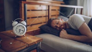 A man sleeping on his side in bed with wooden headboard and bedside table with alarm clock showing time as clocks go back