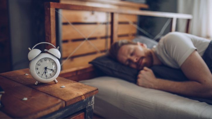 A man sleeping on his side in bed with wooden headboard and bedside table with alarm clock showing time when clocks going back