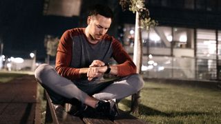 Runner sitting cross-legged on bench at night, checking sports watch