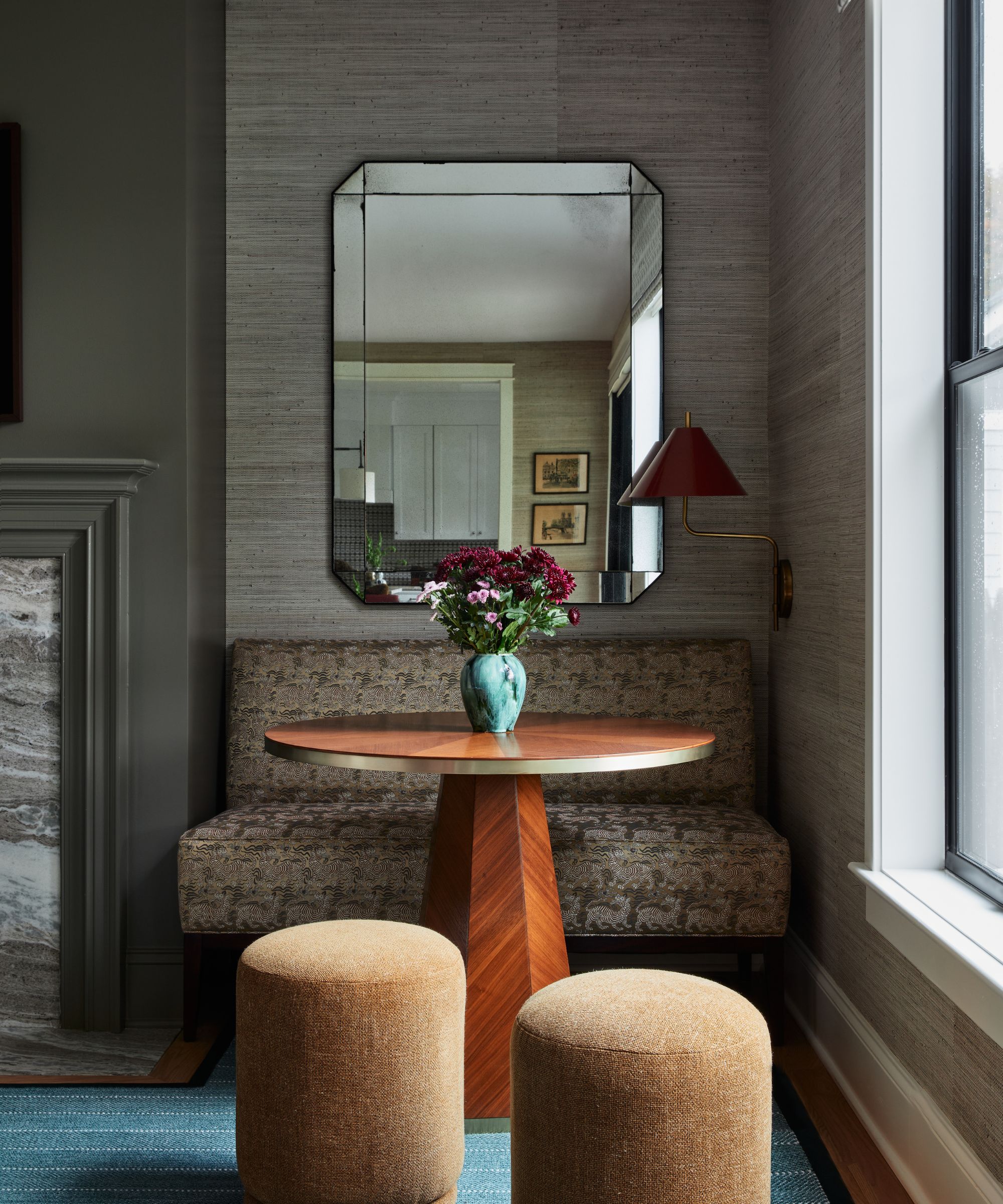A living room with grey grasscloth walls, a nook with a small upholstered banquette, a wooden round table, two brown pouffes, an Art Deco mirror on the wall, a red sconce, and a vase of flowers.