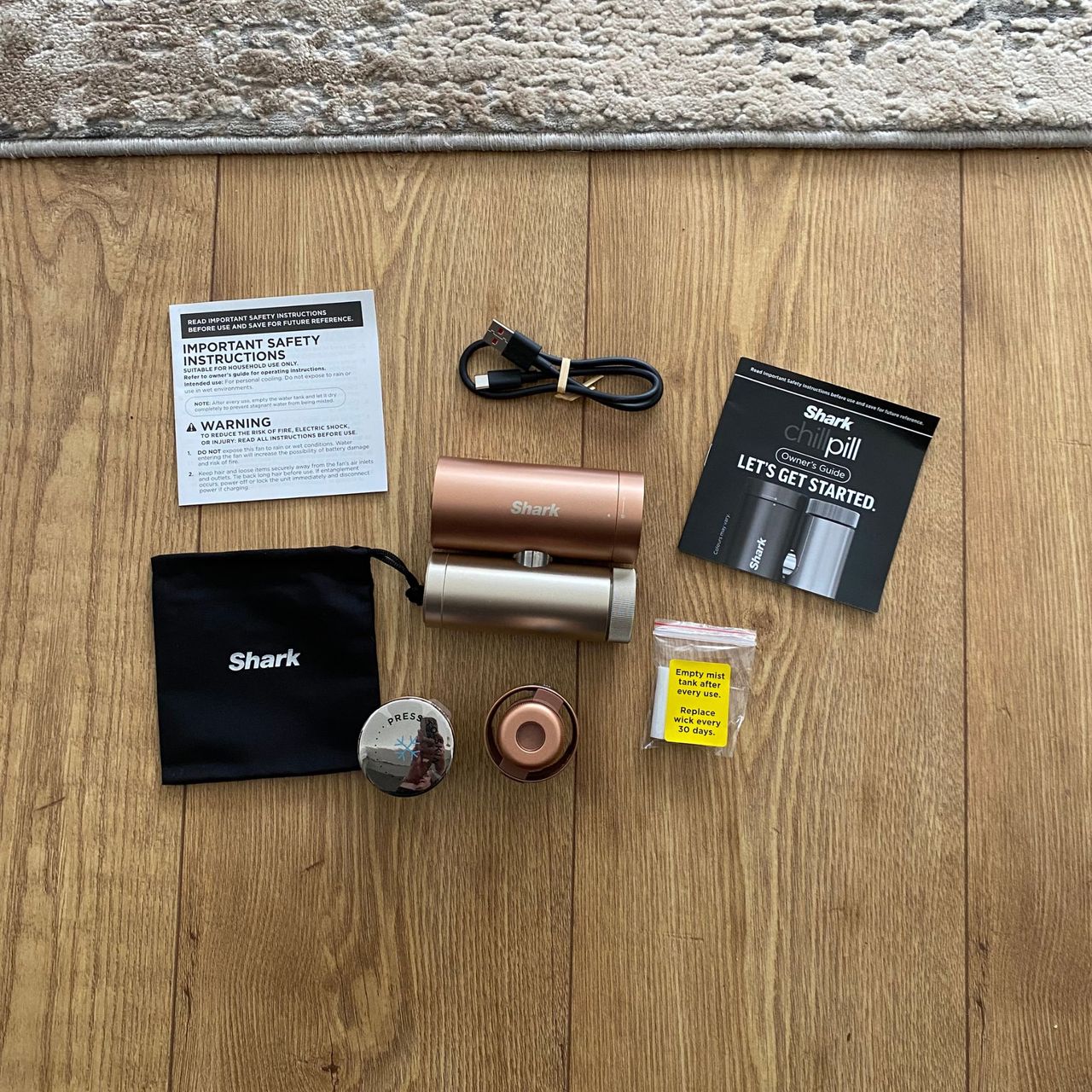 The contents of the Shark ChillPill, including the body, head attachments, storage bag, instructions, charging cable and refill wicks, laid out flat on a wood-look floor