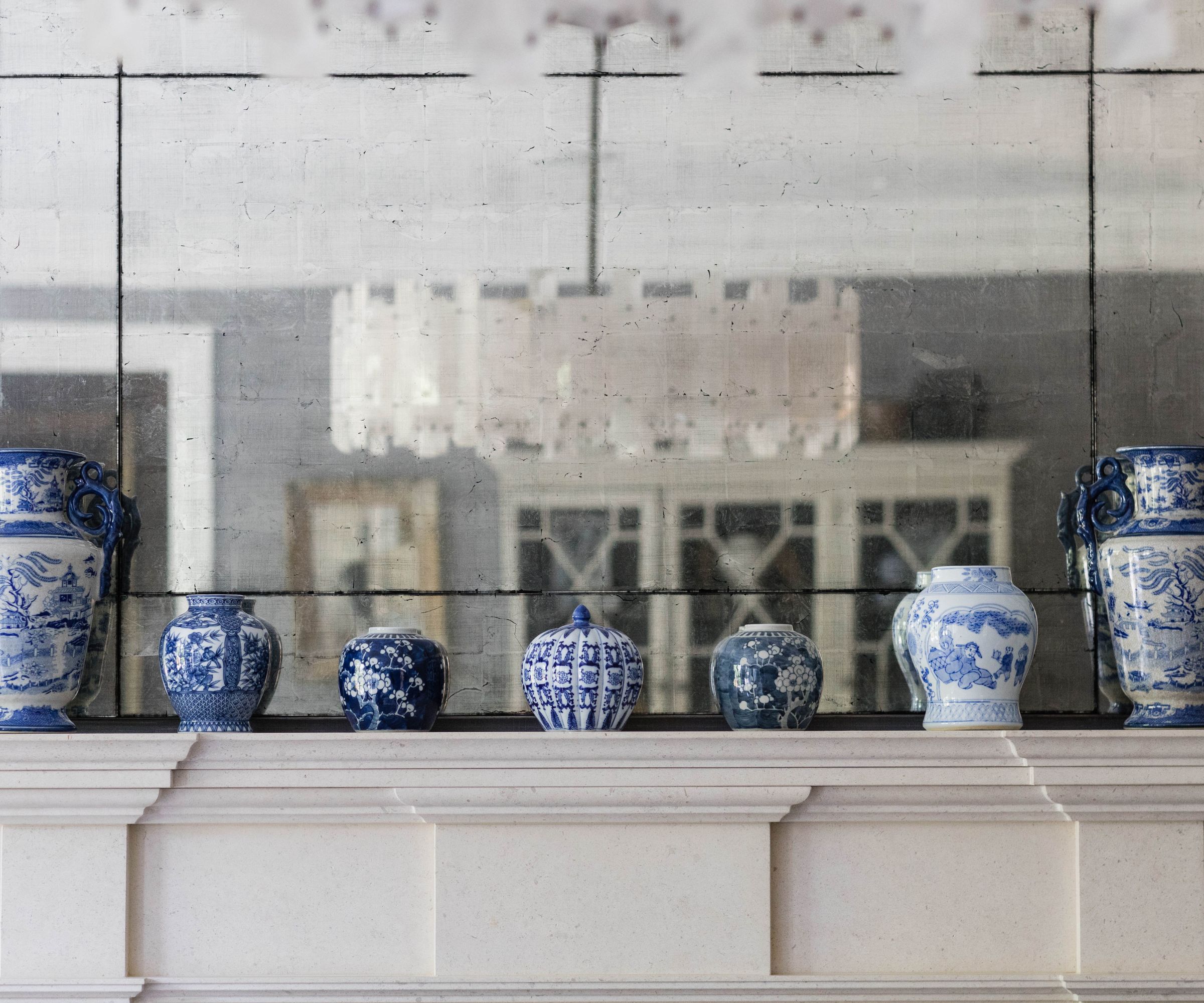 Close up of a collection of blue and white patterned ceramic vases on a mantel. Mirror behind them showing the reflection of a living room
