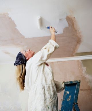 woman wearing white shirt and headscarf on ladder painting ceiling with roller