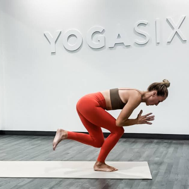 Woman demonstrates yoga pose in yoga studio with words Yoga Six embossed on wall behind her