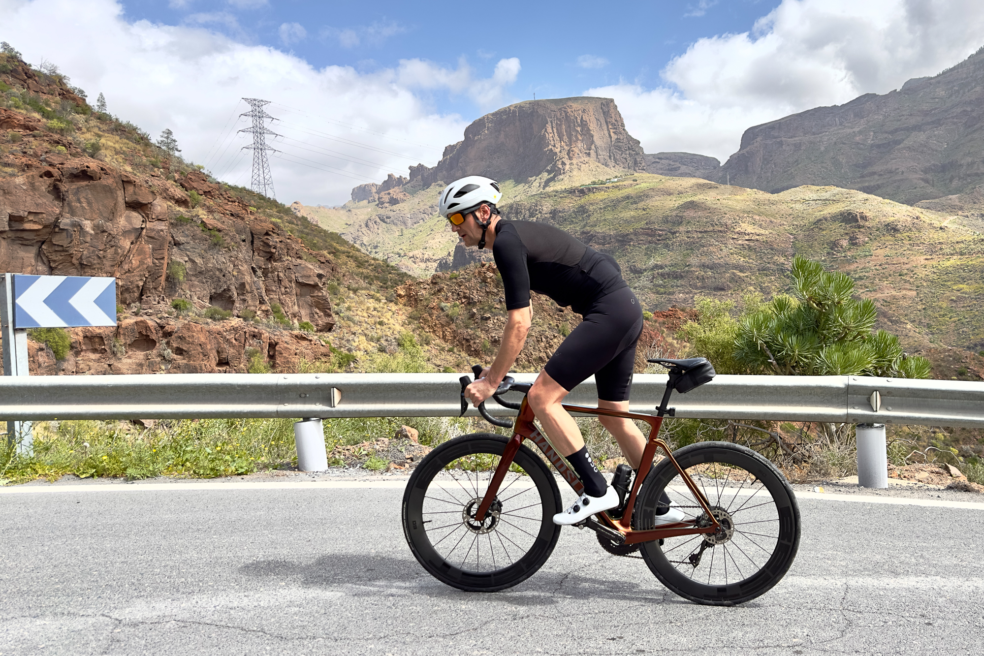 Man wearing a black jersey, black shorts and a white helmet riding an orange road bike up a hill out of the saddle