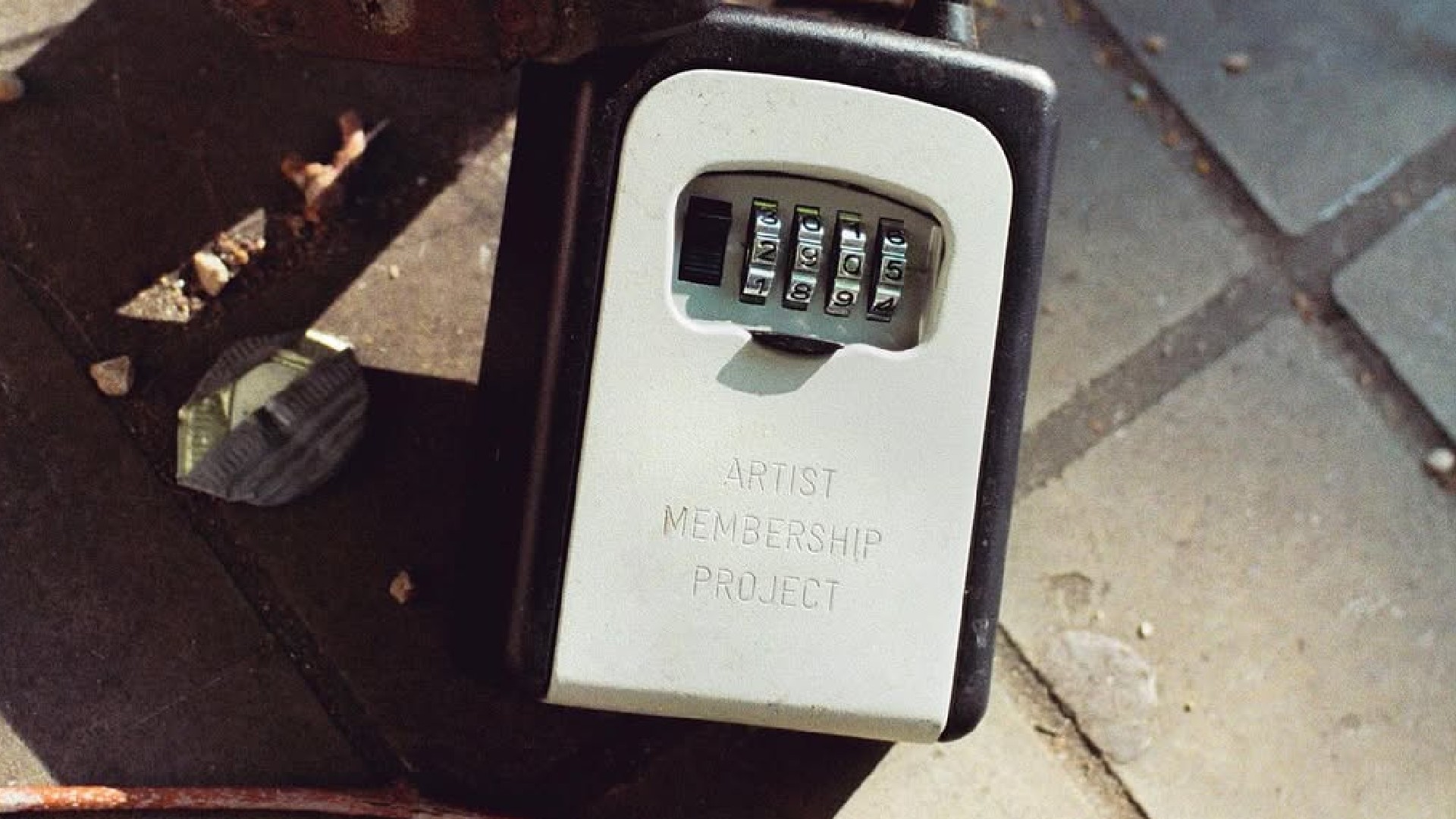 A close-up shot of a key safe box on the ground with a four-digit combination lock, inscribed with the text &quot;ARTIST MEMBERSHIP PROJECT&quot; on its pale gray face.