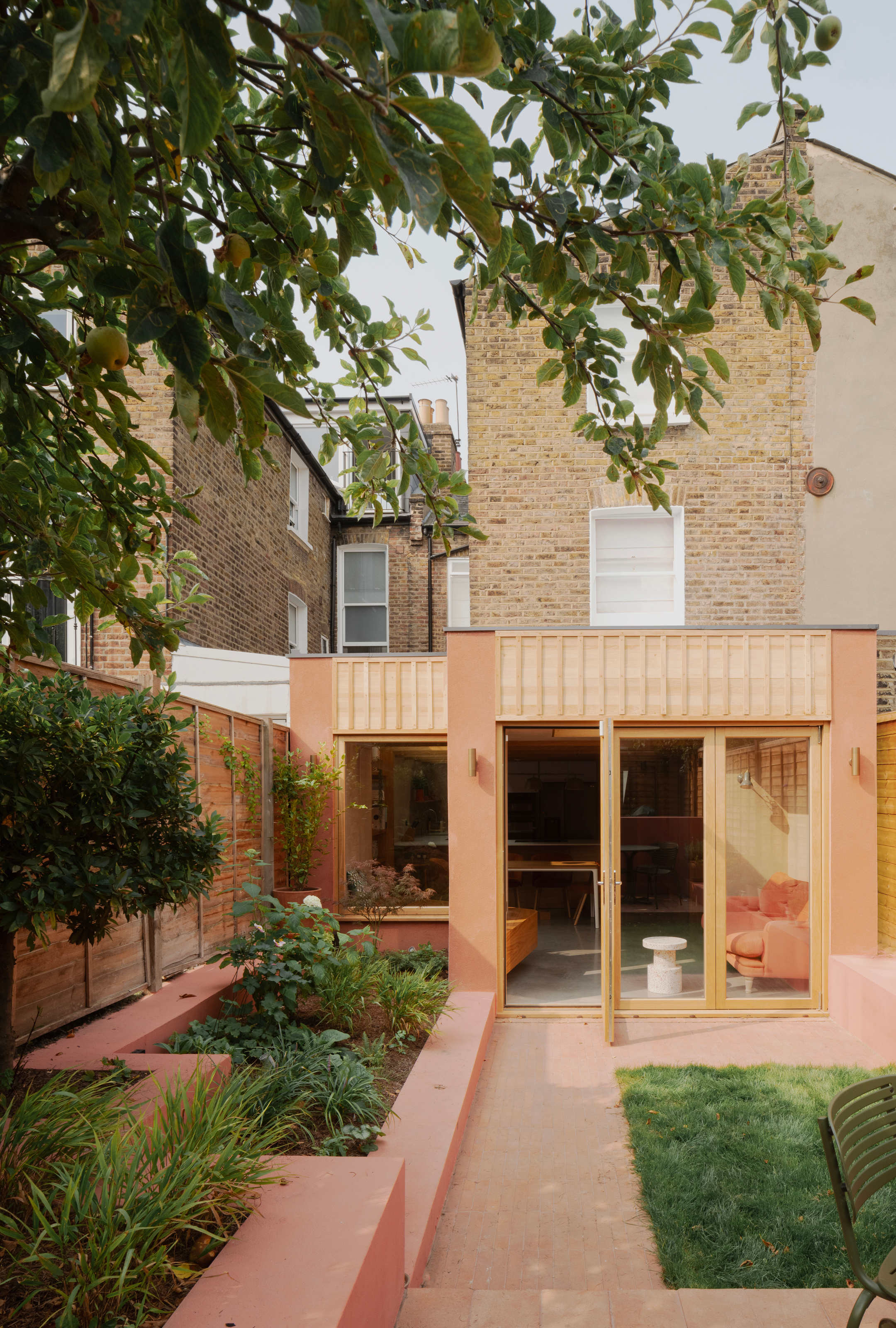 Extension with apricot polished concrete and wood-clad finish with pink-hued garden beds filled with plants and a small square of grass in the center with a trees' leaves hanging just in shot