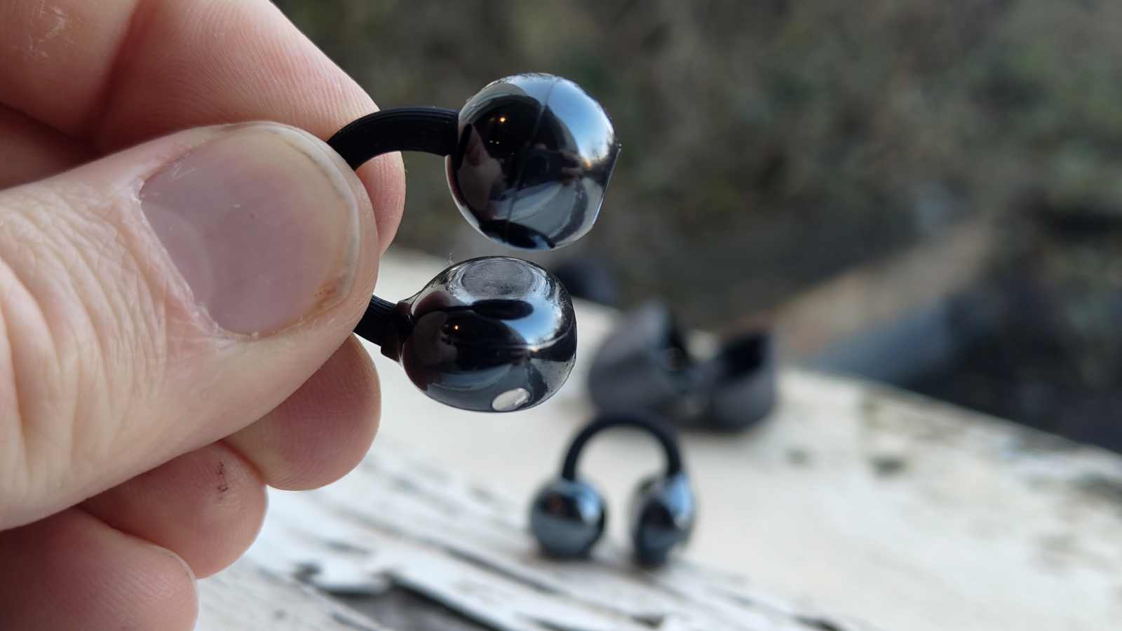 The Huawei FreeClip 2, with one bud between two fingers and the other with the case on a shelf behind.