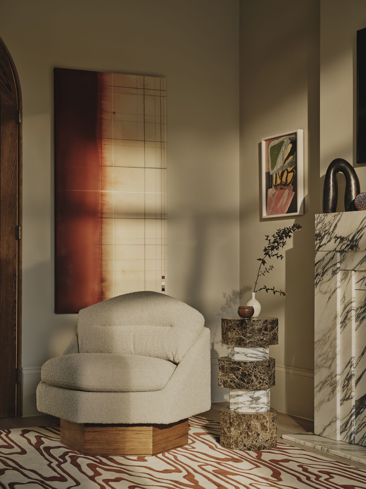 Image of the corner of a living room with a marble fireplace in view. There is an off-white geometric accent chair with a wooden base on top of a white and terracotta red rug that has a wavy line pattern. Beside the chair is a stacked marble accent table.