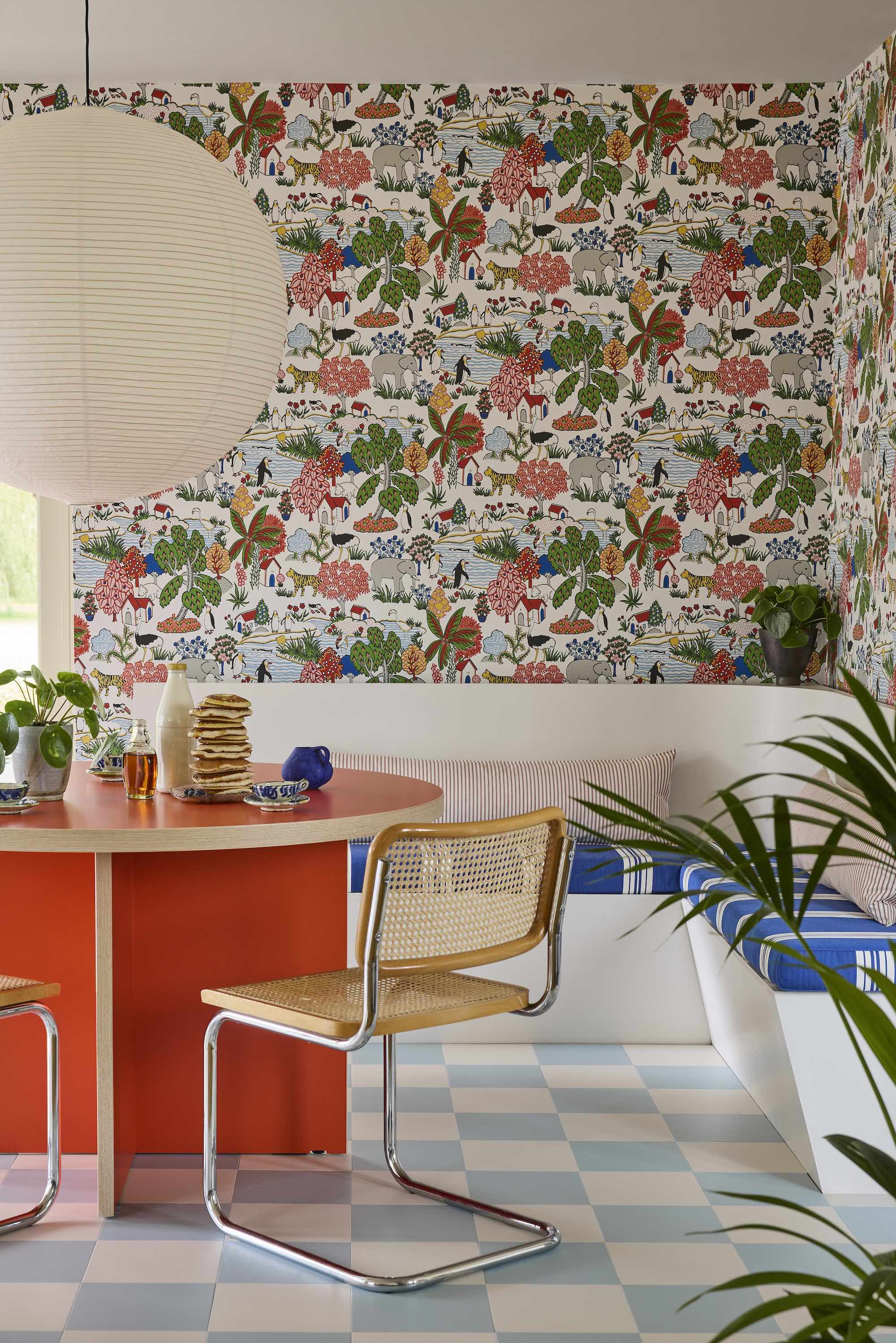 Dining are with an orange circle dining table to the left and a white, corner bench seat to the right. A large, white paper light hangs above the table and the walls are covered in a vibrant patterned wallpaper.