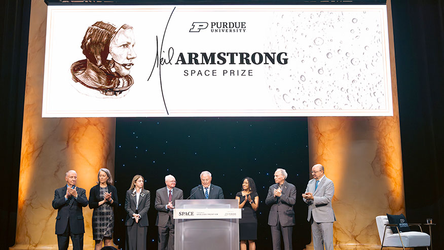 eight men and women in evening wear stand together on stage under a large white sign for Purdue University's Neil Armstrong Space Prize