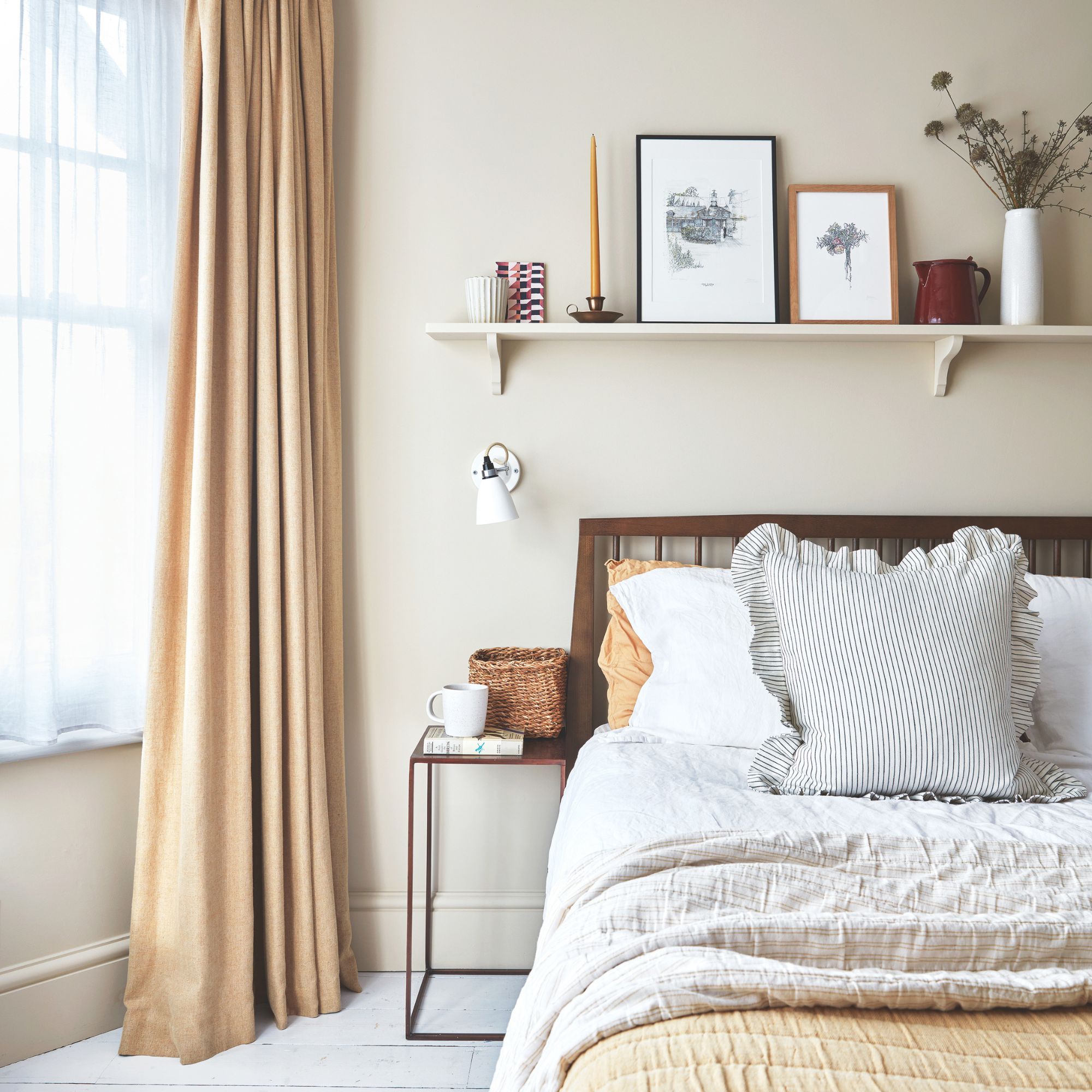 Beige painted bedroom with a window and curtains to the left hand side and a bed with a shelf above it to the right