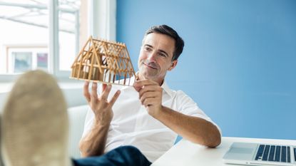 A man smiles pensively as he looks at a model of a house that he is holding.