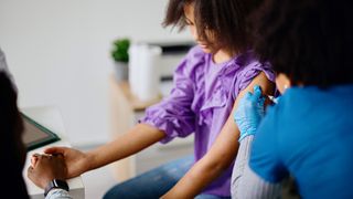 A teen girl receives an HPV vaccine while sat in a doctor's office. She is holding her mother's hand.