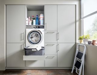 grey utility room Schüller, with pull out-drawer for laundry basket, a washing machine, and cleaning products in a shelf, with a wooden floor and a stepladder and window to the right
