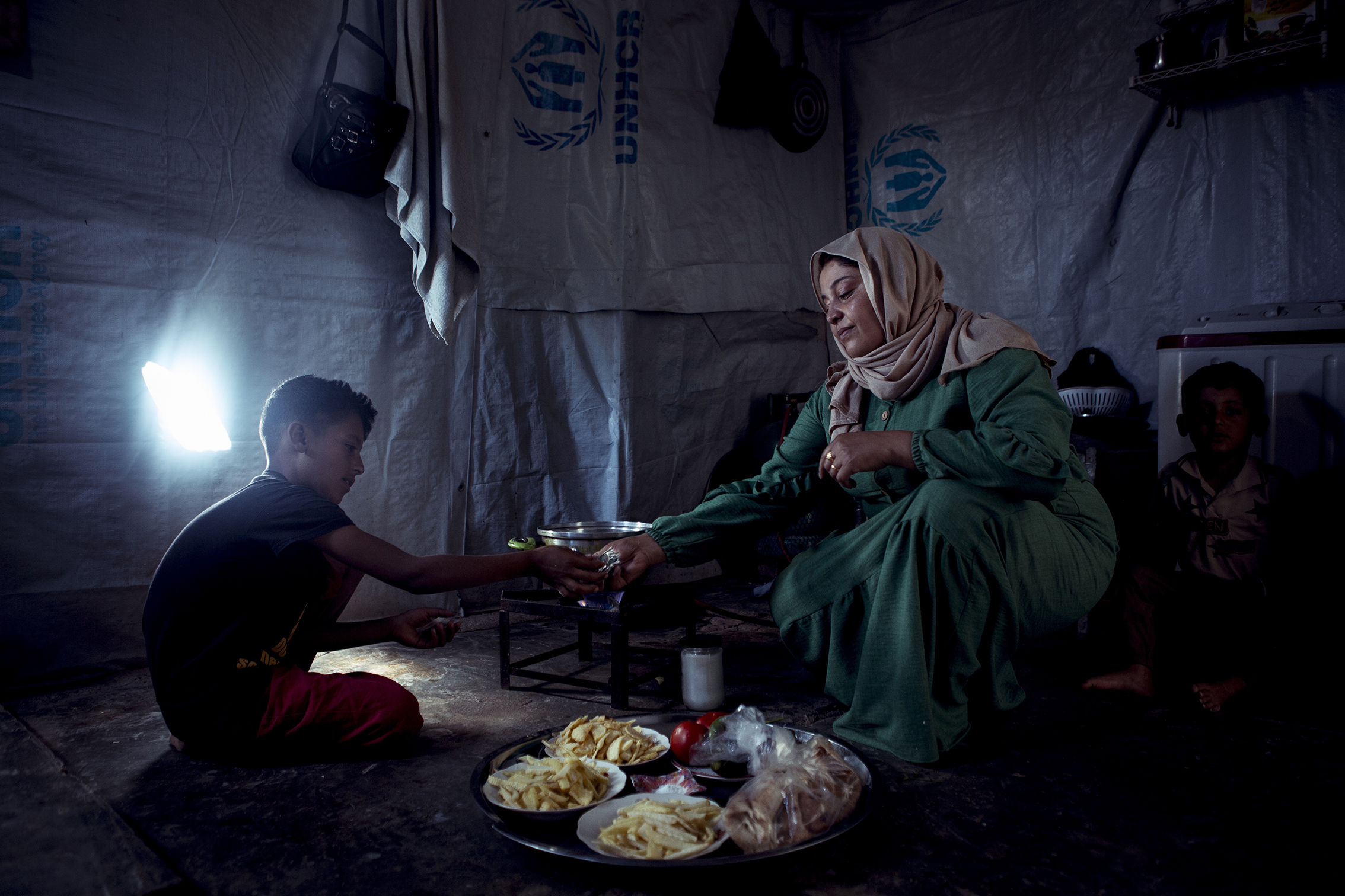 A woman in a green dress and headscarf shares food with a child in a dimly lit tent with UNHCR logos. The mood is intimate and caring. Plates of food are on the floor