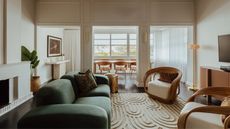 Image of a white living room with crown molding on the walls. There is a large, overstuffed, emerald green sofa and two accent chairs made of fluted wood and a white square cushion. There is a rug that has a geometric design.