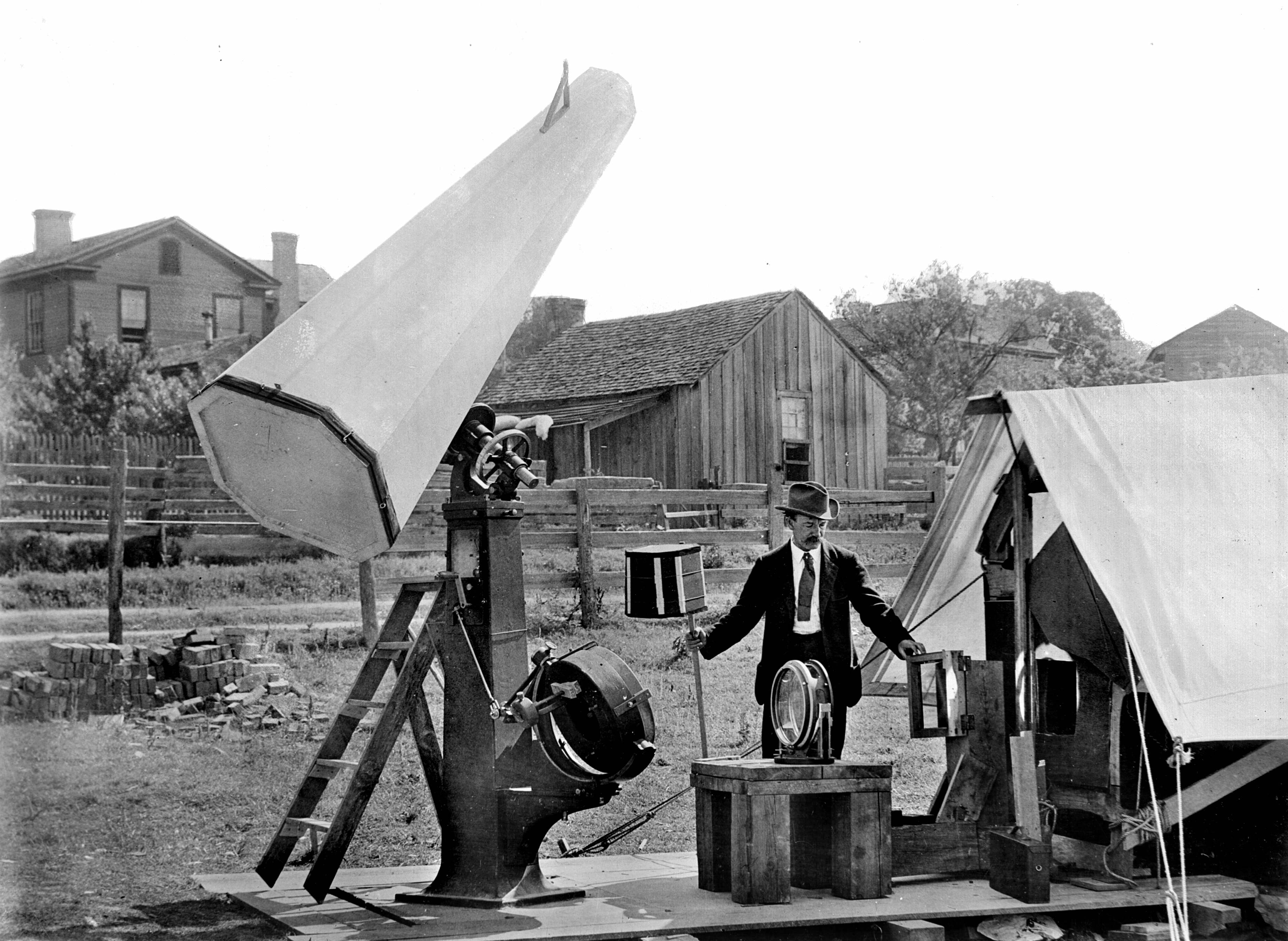 A man stands next to a large telescope and a shelter tent which housed a photographic room. He is wearing a suit and hat.