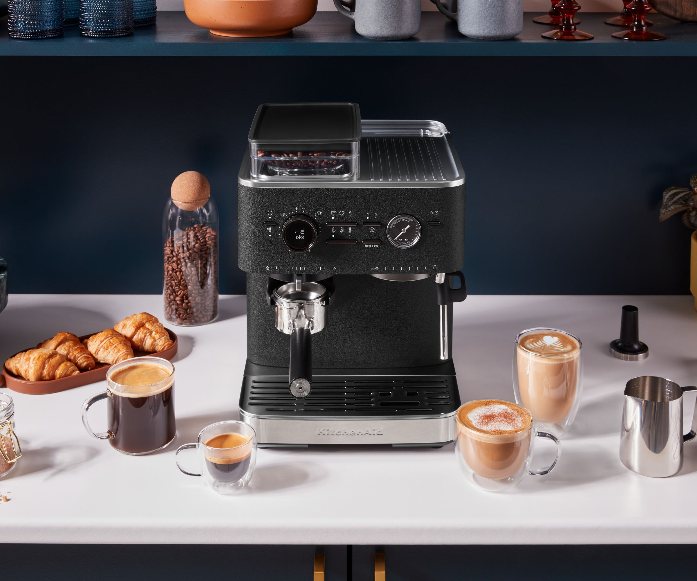A black countertop espresso machine displayed on a white kitchen worktop, styled with coffee beans, croissants, espresso cups, cappuccinos and a milk jug, set against dark blue cabinetry for a modern coffee station setup.