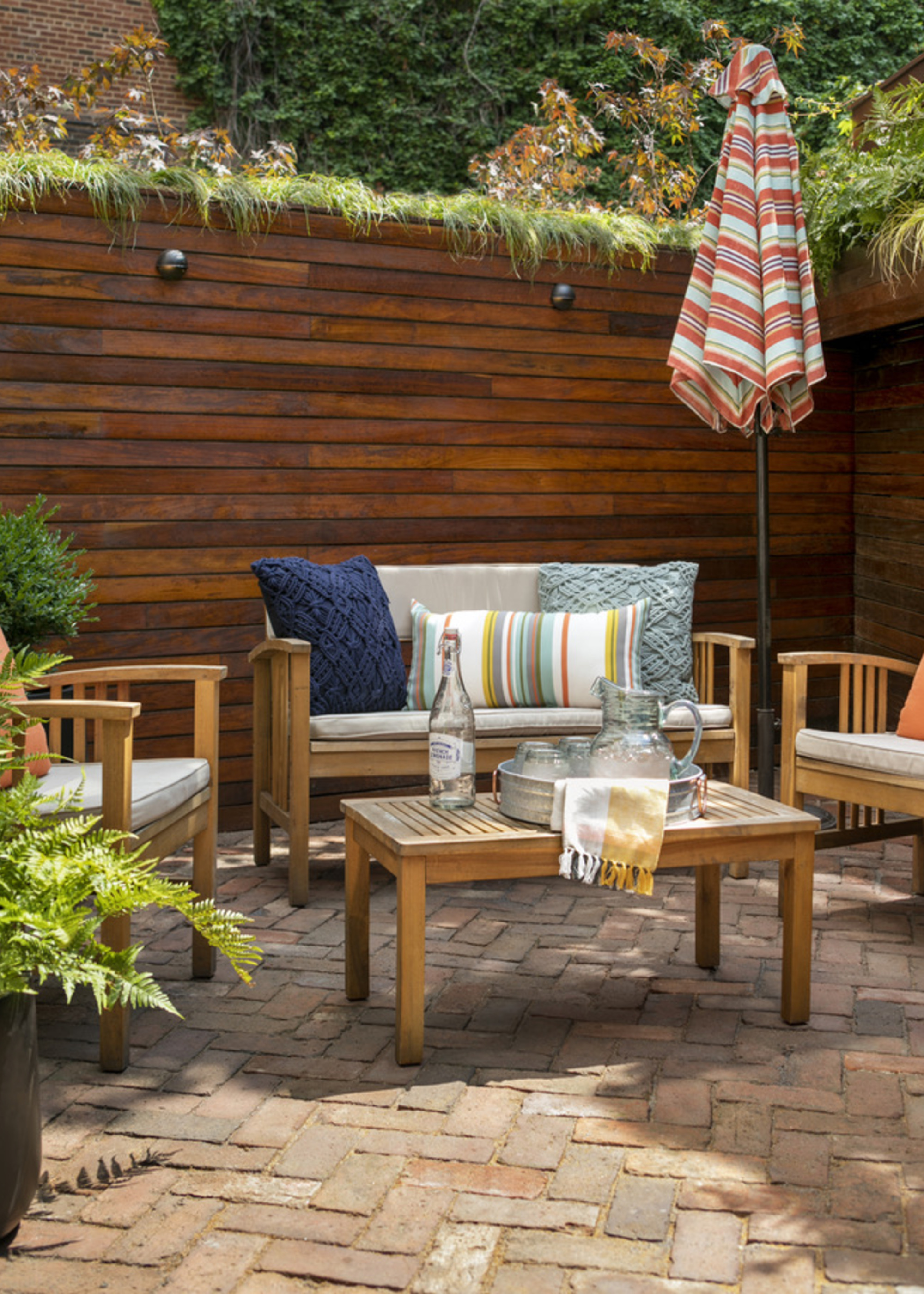 A patio with brick flooring, wooden furniture, a tall, modern slatted fence, and a striped parasol