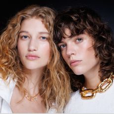 two models backstage at roberto cavalli spring 2025 fashion show wearing white shirts and gold necklaces with clean makeup
