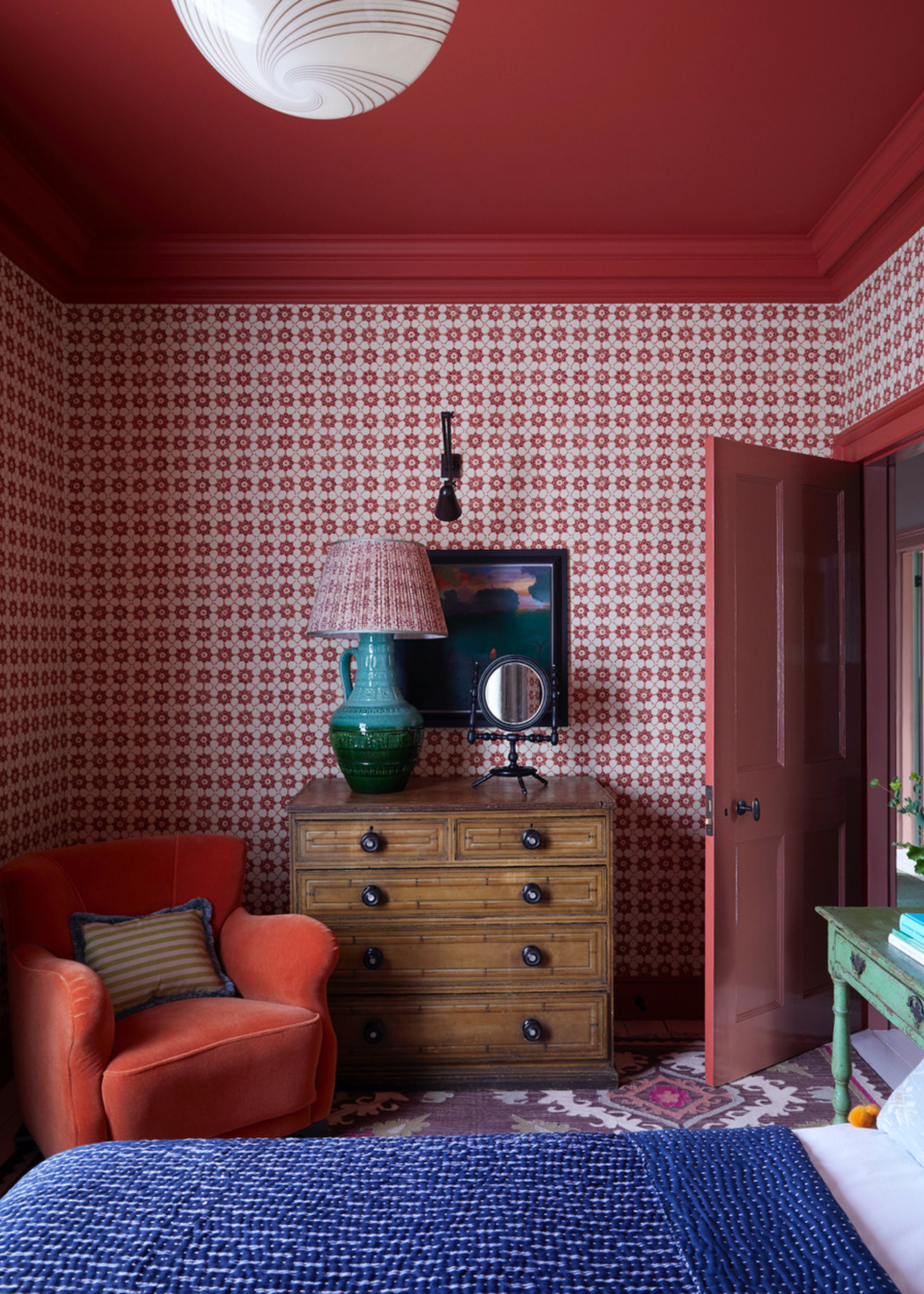 bedroom with red ceiling, red patterned wallpaper, blue bedcover and white sheets, patterned carpet, red armchair with striped cushion, and timber chest of drawers with mirror and lamp styled on top with artwork hung on the wall above