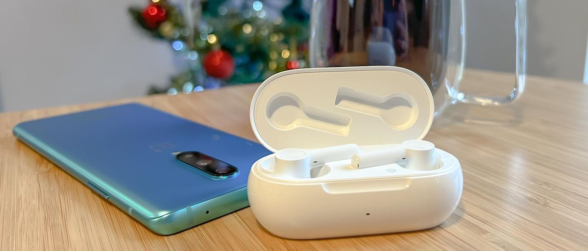 The OnePlus Buds Z2 in its charging case, next to a OnePlus 8 phone and a glass mug of coffee, on a wooden table with a Christmas tree in the background