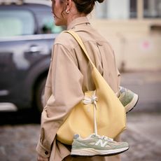 Woman wears tan button down while carrying a large yellow handbag with green New Balance sneakers hanging off of the handbag. 