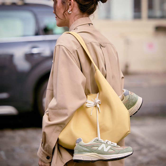 Woman wears tan button down while carrying a large yellow handbag with green New Balance sneakers hanging off of the handbag. 