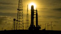The silhouette of a rocket is pictured at a launch pad with the sun behind at sunset. The sky is golden.