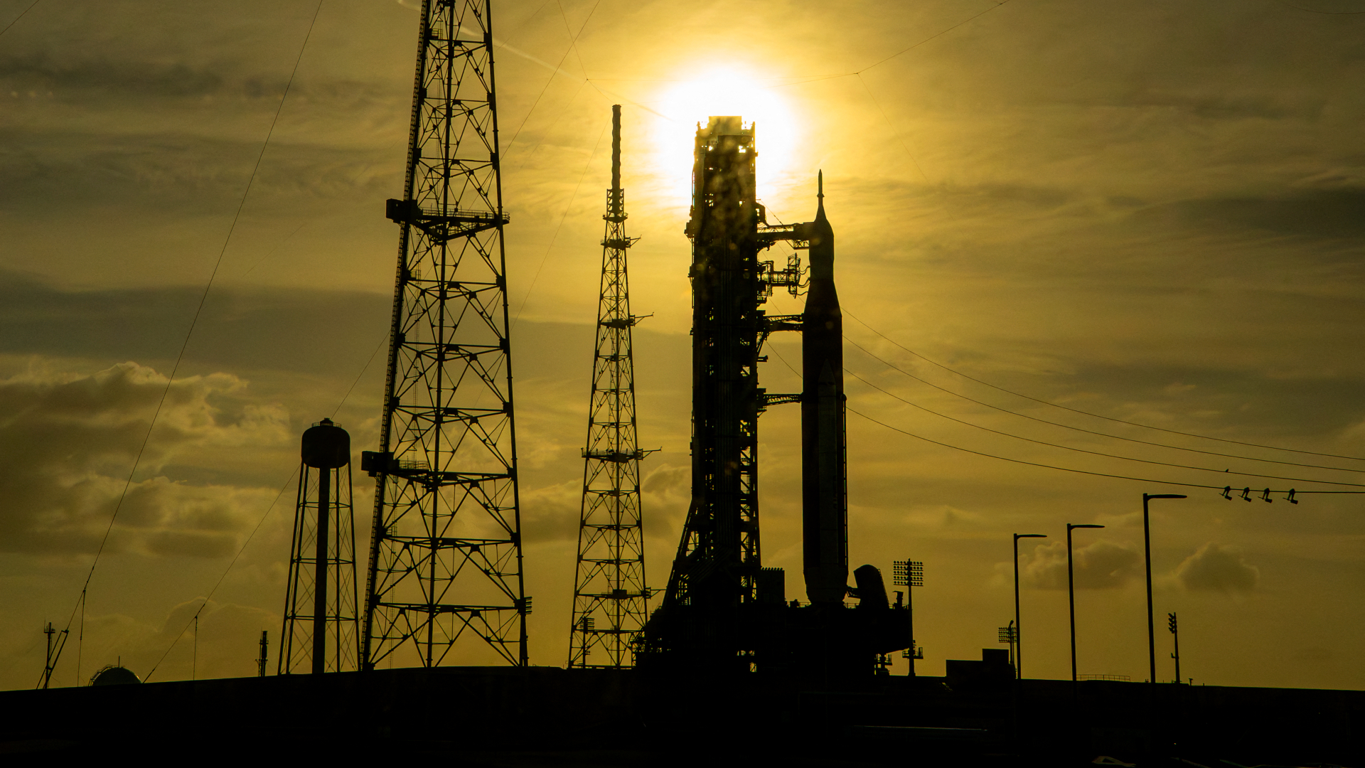 The silhouette of a rocket is pictured at a launch pad with the sun behind at sunset. The sky is golden.