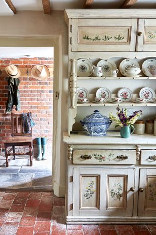 a traditional white freestanding dresser in a country kitchen with red tile floor, a wooden chair, and welly boots in the background