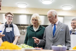 Queen Camilla wearing a green coat and King Charles wearing a gray suit and pointing at food in a kitchen