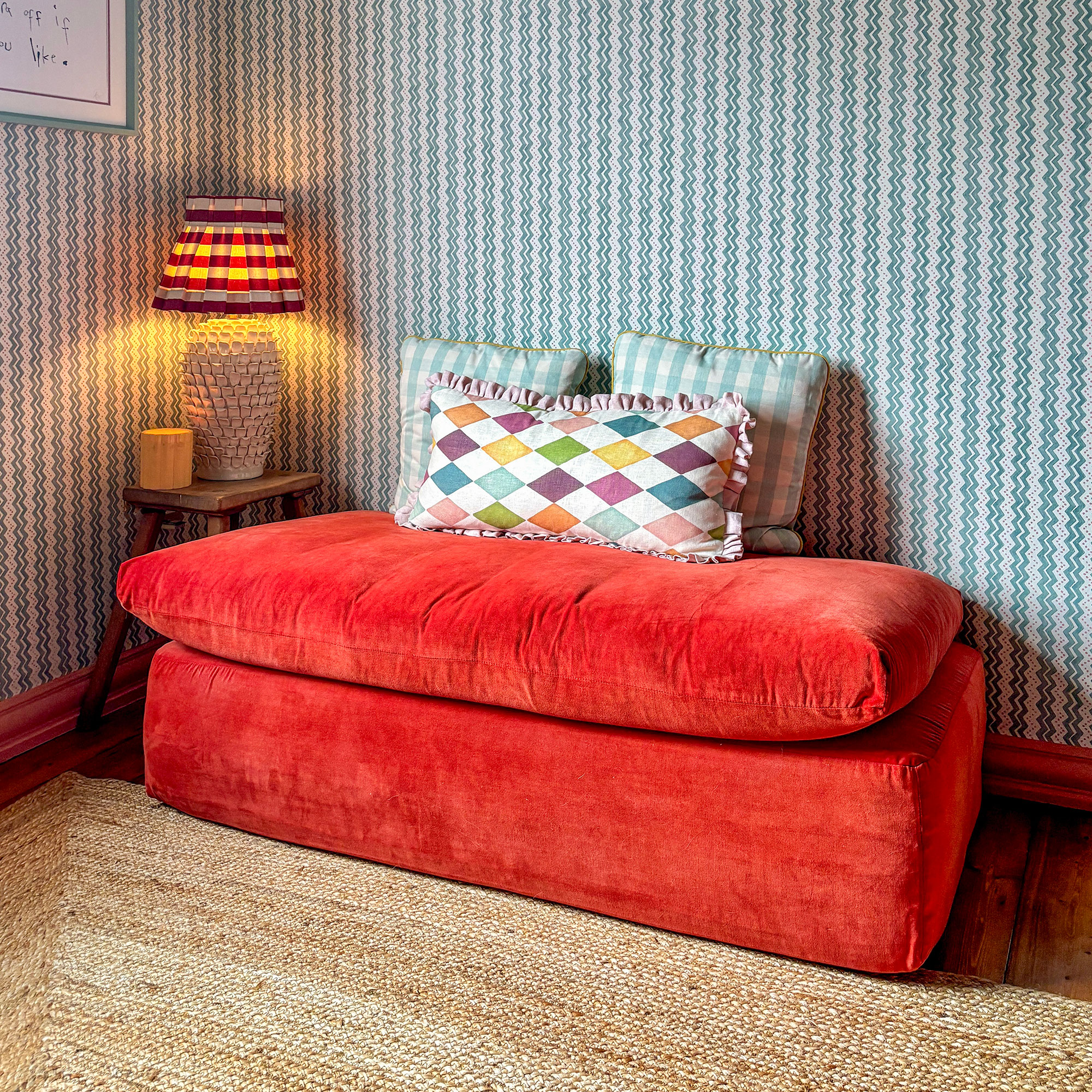 Orange red fabric bench topped with patterned cushions, on woven rug and in front of blue and white striped wallpaper