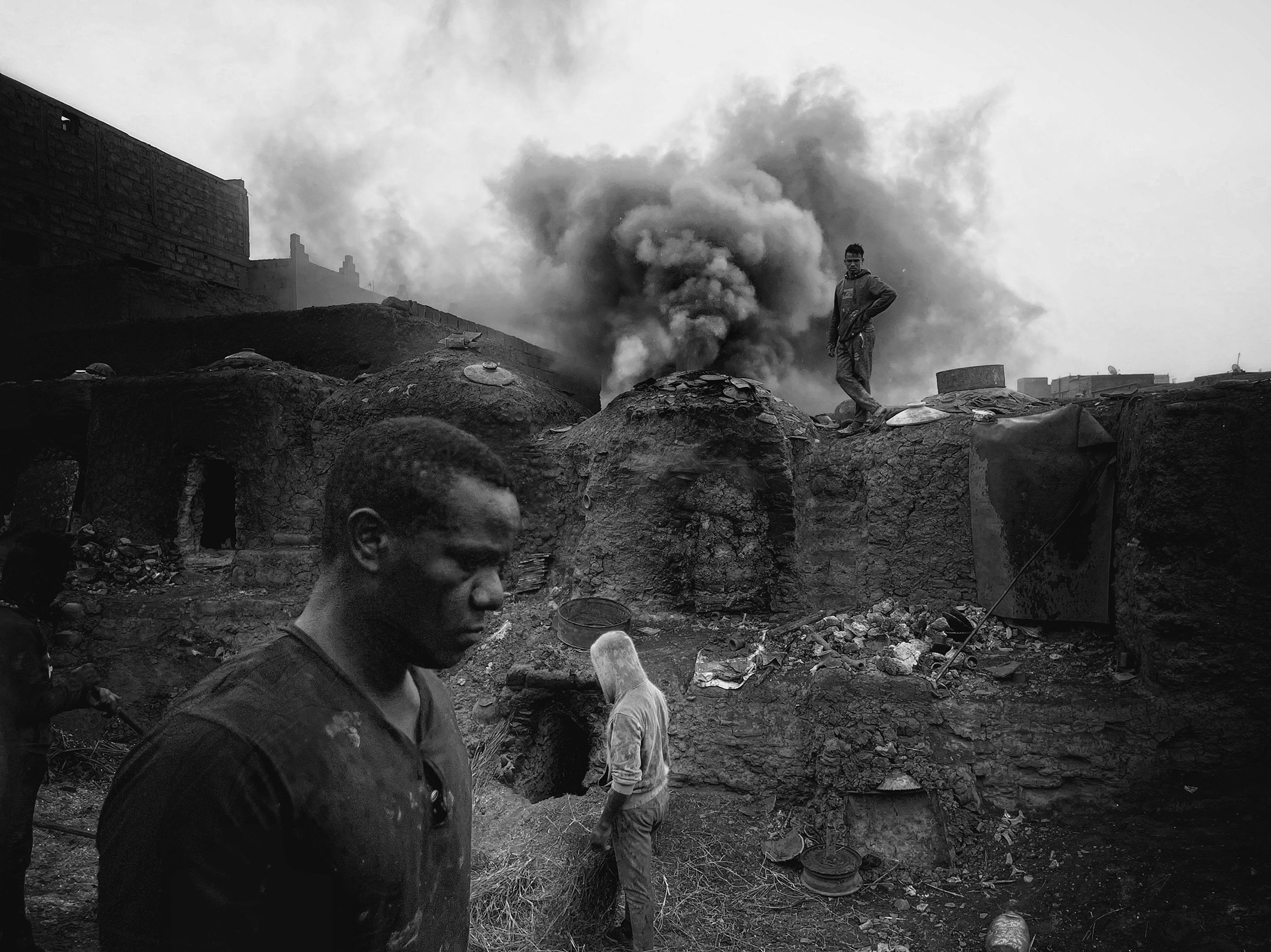 A smoky scene in a desolate environment, featuring men navigating debris and ruins amid billowing black smoke