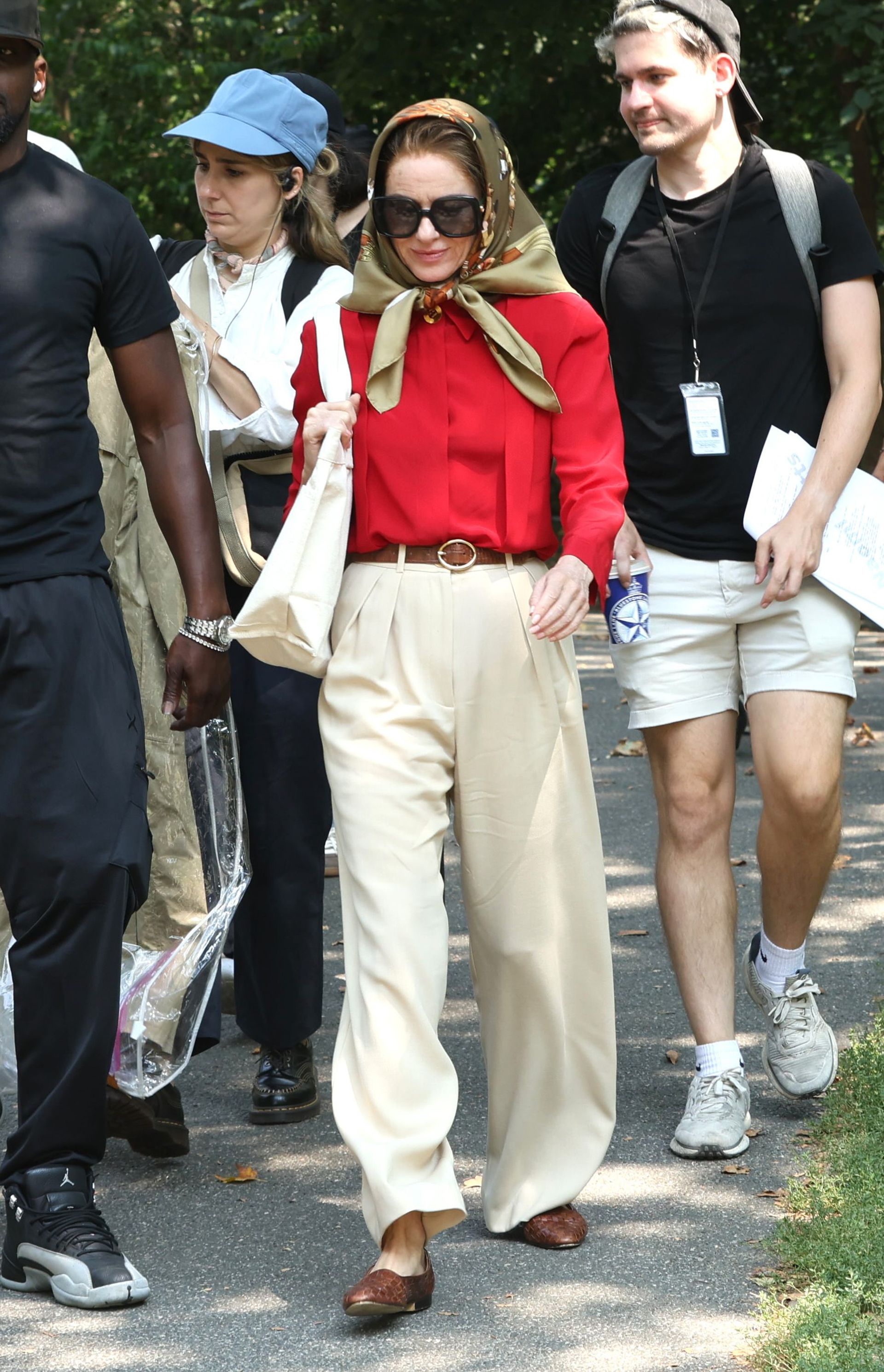 Naomi watts transforms into jackie kennedy wearing a headscarf, large sunglasses, a red blouse, brown belt, beige wide-leg pants, and brown woven leather ballet flats.