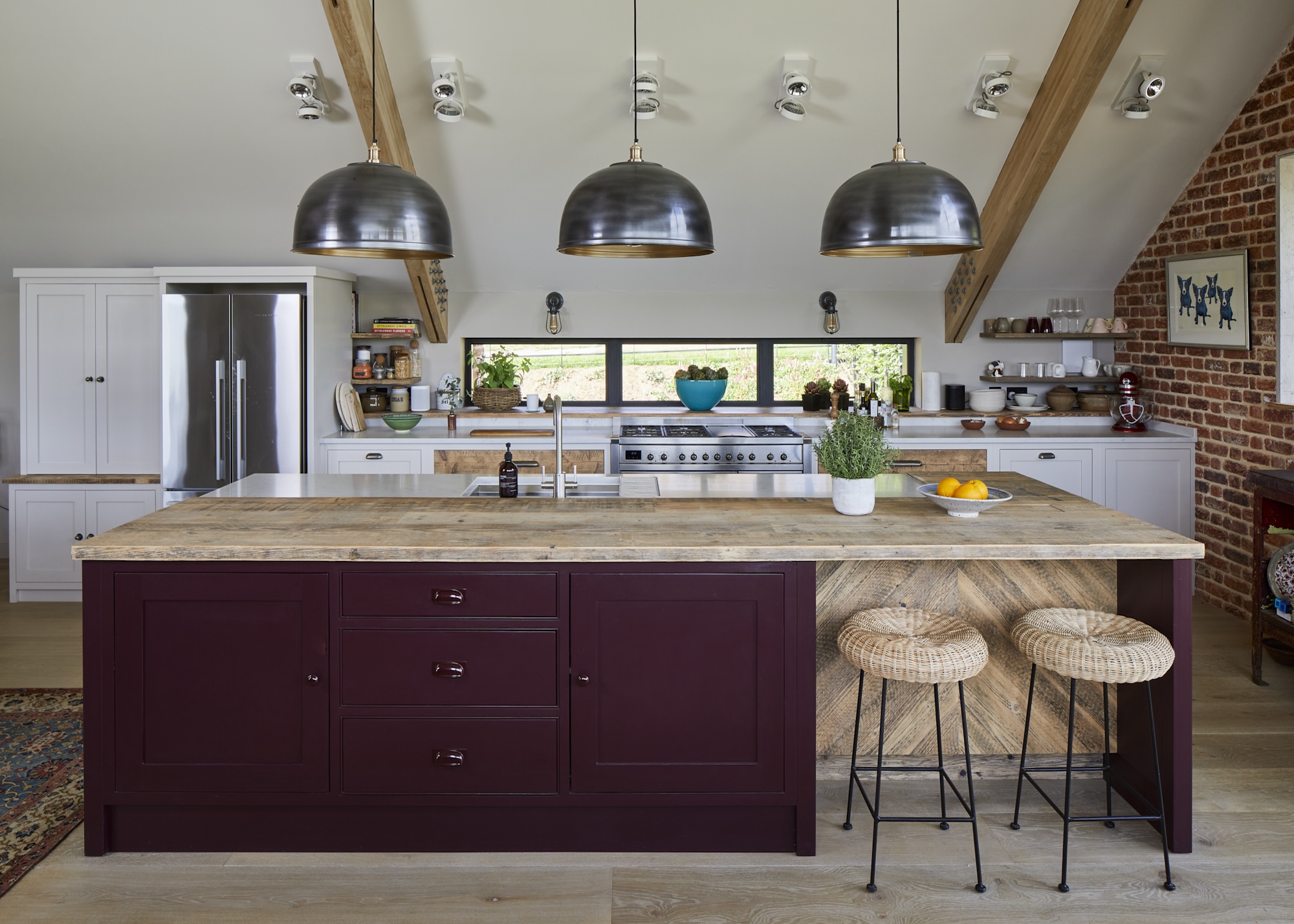 A large kitchen island in the middle of a large kitchen. Above the island are three pendant lights hanging down as well as two bar stools beside the island. There is a view of the stove as well as fridge and other key elements within the kitchen