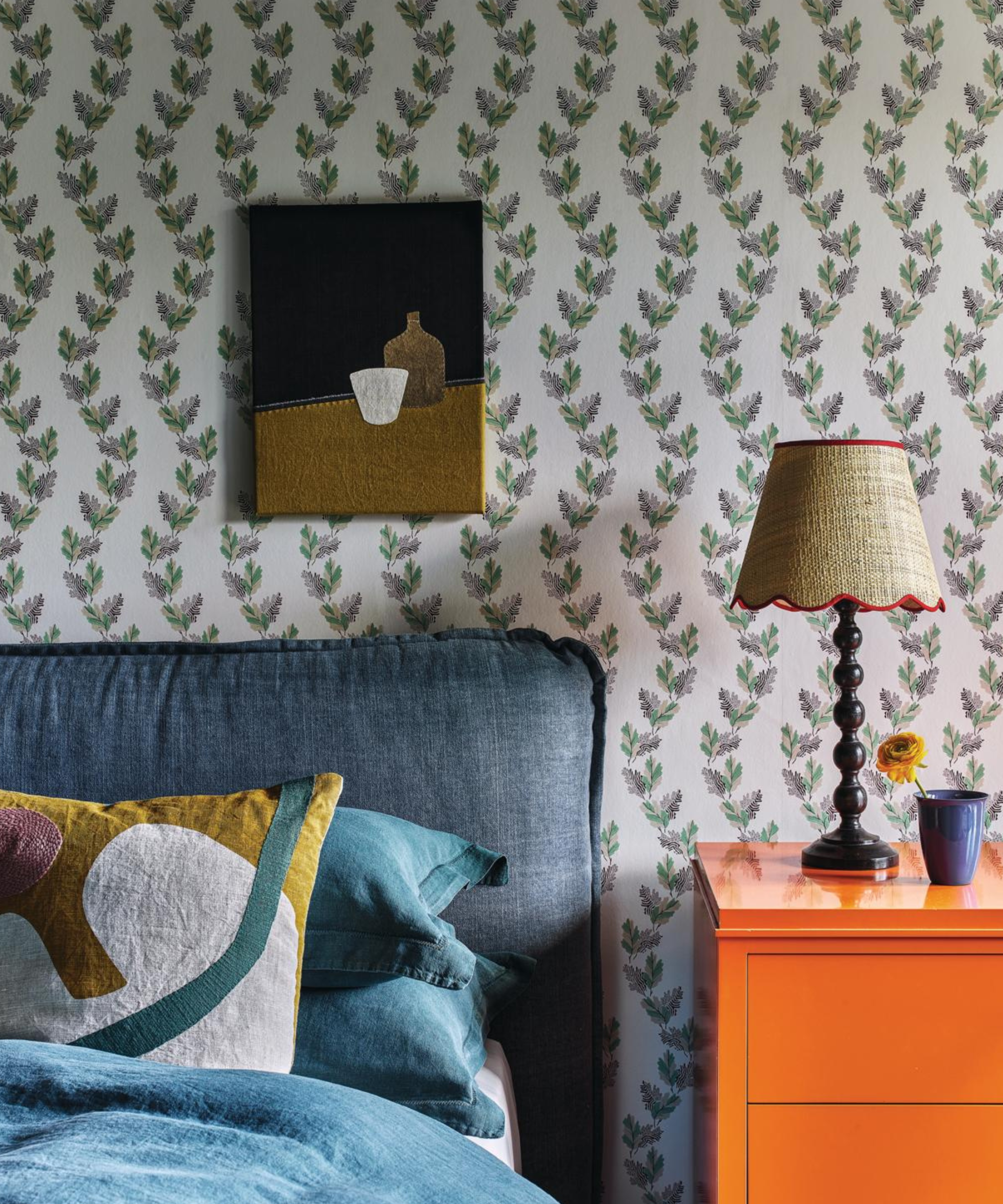 a bedroom with floral vine wallpaper, a bed with a blue headboard, and a bright orange side table
