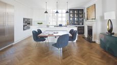 A white and blue kitchen with wooden flooring. In the centre is a large dining table with blue velvet chairs, with gold legs. Behind is a white kitchen island with blue pendant lamps above, and white and blue cabinets behind that. To the left is a large silver fridge freezer, and to the right is a white mantelpiece with framed artwork above, a green sideboard, and a black and gold lamp with a white lampshade.